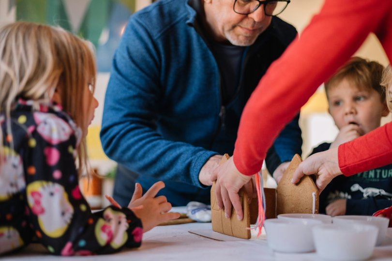 Photo avec deux personnes faisant une maison en pain d’épice et trois enfants les regardant